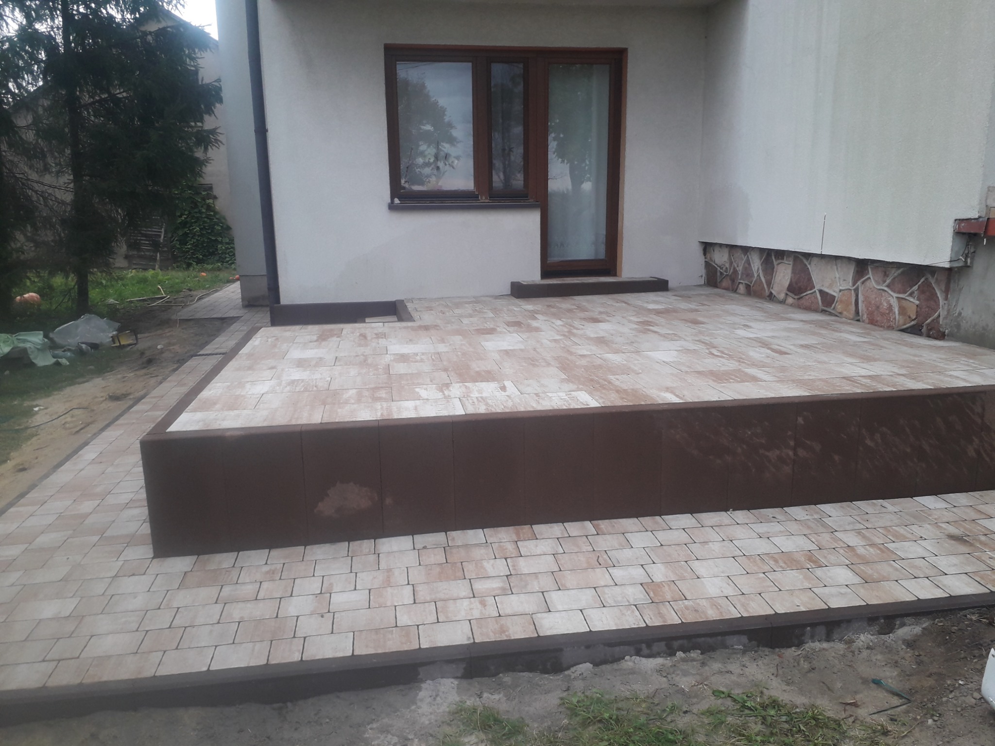 Taras z jasnej kostki brukowej z brązowym obrzeżem i podwyższeniem, widok na okno i drzwi balkonowe budynku.