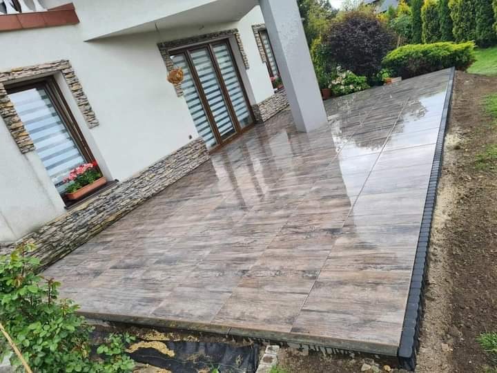 Wyłożony płytkami taras przy domu z oknami ozdobionymi kamienną okładziną i skrzynką z kwiatami, widoczne odbicia w mokrej powierzchni płytek.