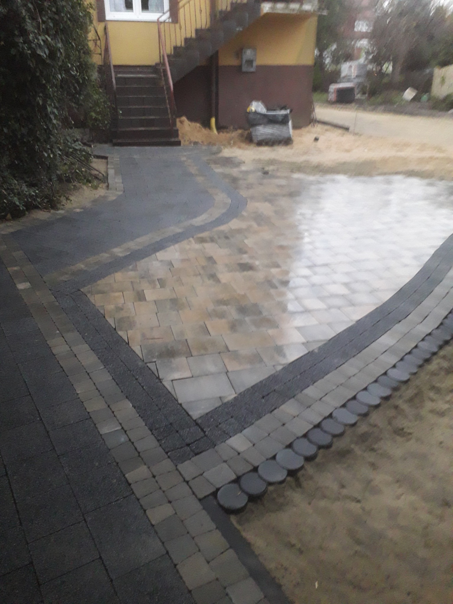 Ułożona kostka brukowa w różnych odcieniach szarości i beżu tworząca ścieżkę i plac przed budynkiem z zewnętrznymi schodami, widoczne palety z kostką w tle.