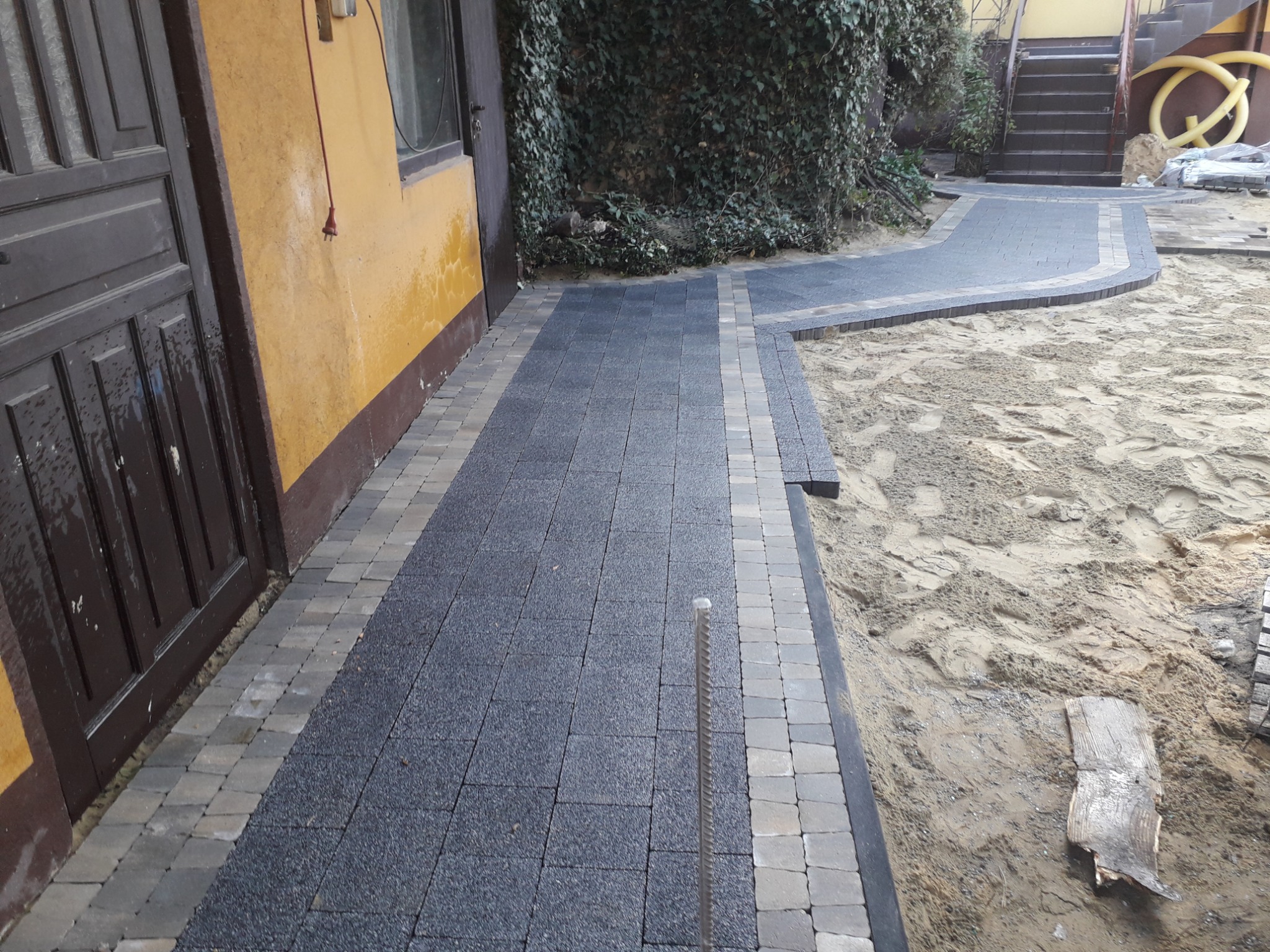 Ułożona ścieżka z ciemnej kostki brukowej z jasnym obramowaniem, prowadząca wzdłuż ściany budynku z brązowymi drzwiami i schodami w tle, teren wokół w trakcie prac wykończeniowych.
