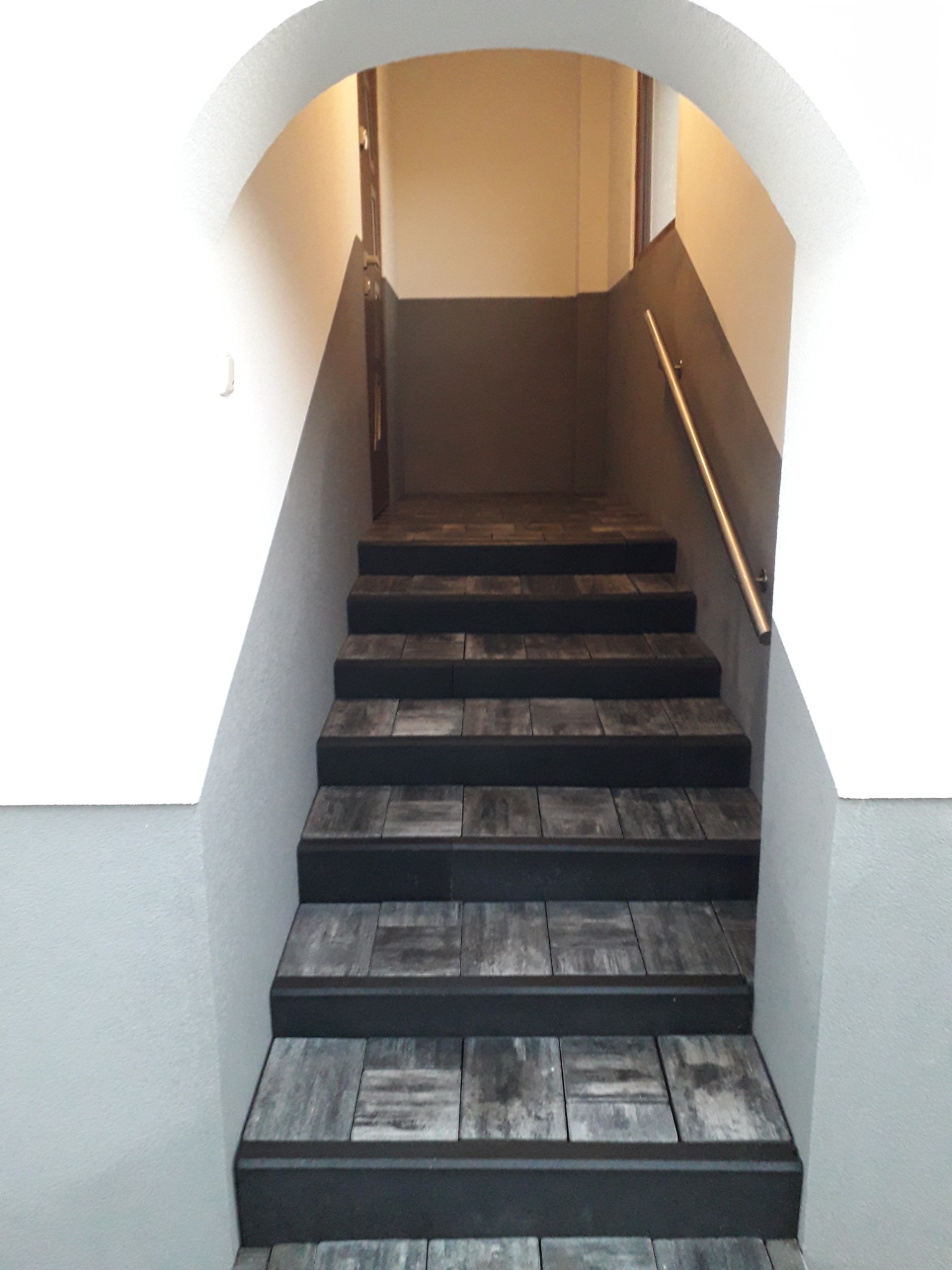 Schody wyłożone szarą kostką brukową wewnątrz budynku, z metalową poręczą po prawej stronie i łukowatym wejściem.