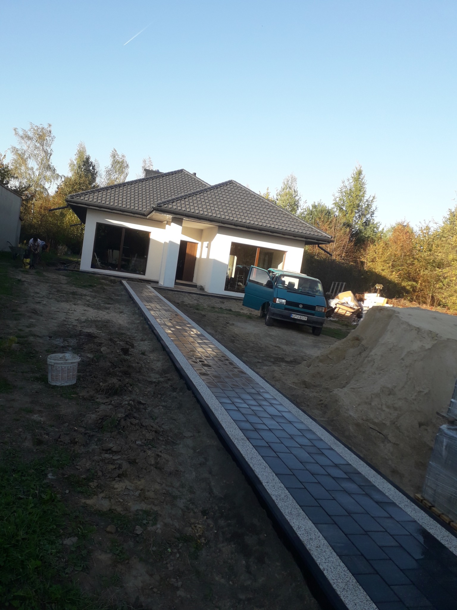 Nowo ułożony, prosty chodnik z ciemnej kostki brukowej z jasnoszarymi obrzeżami prowadzi do nowoczesnego, parterowego domu z ciemnym dachem; w tle zaparkowany turkusowy bus i usypana pryzma piasku.