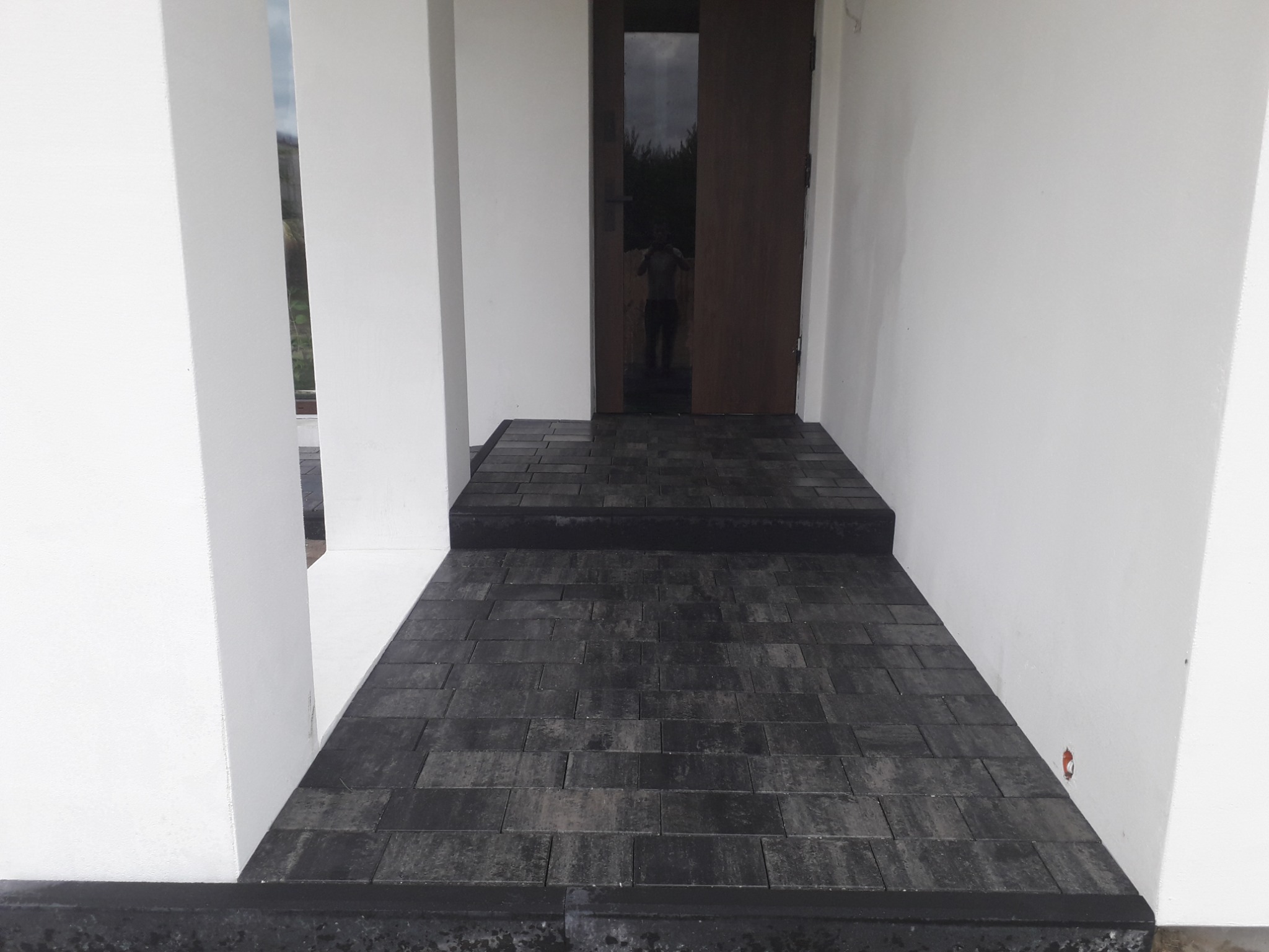 Ciemnoszara kostka brukowa prowadząca do wejścia domu z białymi filarami i drewnianymi drzwiami z odblaskiem sylwetki.