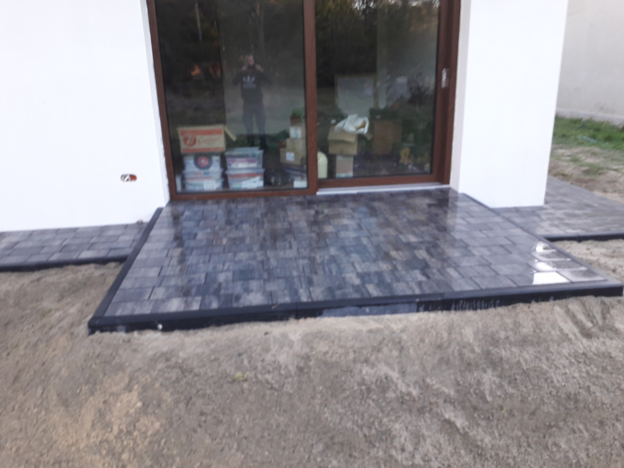 Nowo ułożona, mokra, szara kostka brukowa przed wejściem do domu z brązowymi drzwiami, widoczny piasek i odbicie sylwetki w szybie.