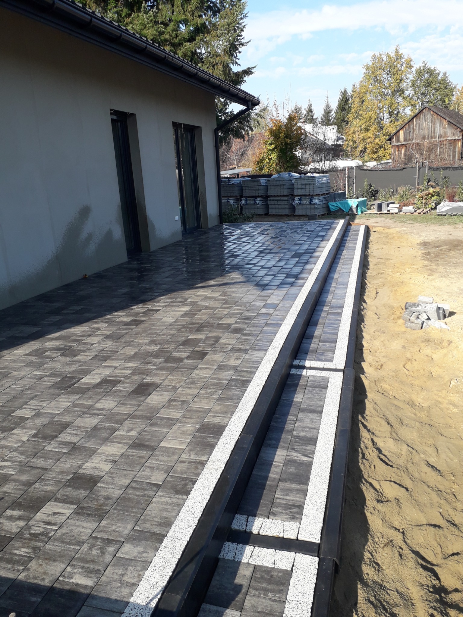 Ułożona, grafitowa kostka brukowa z białymi obrzeżami na tarasie przylegającym do budynku, z widoczną stertą nieużytych kostek w tle.