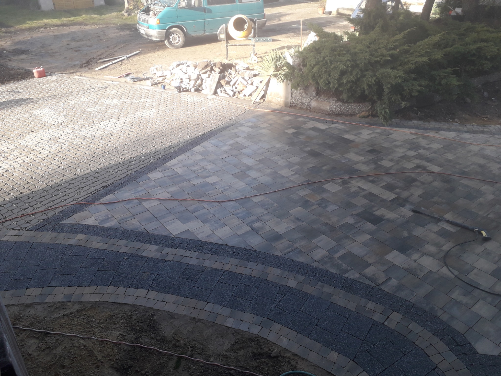 Fragment nowo ułożonego podjazdu z kostki brukowej w odcieniach szarości, z widocznymi elementami dekoracyjnymi w postaci ciemniejszych pasów i obrzeży, na tle zaparkowanego busa i sterty kostki.