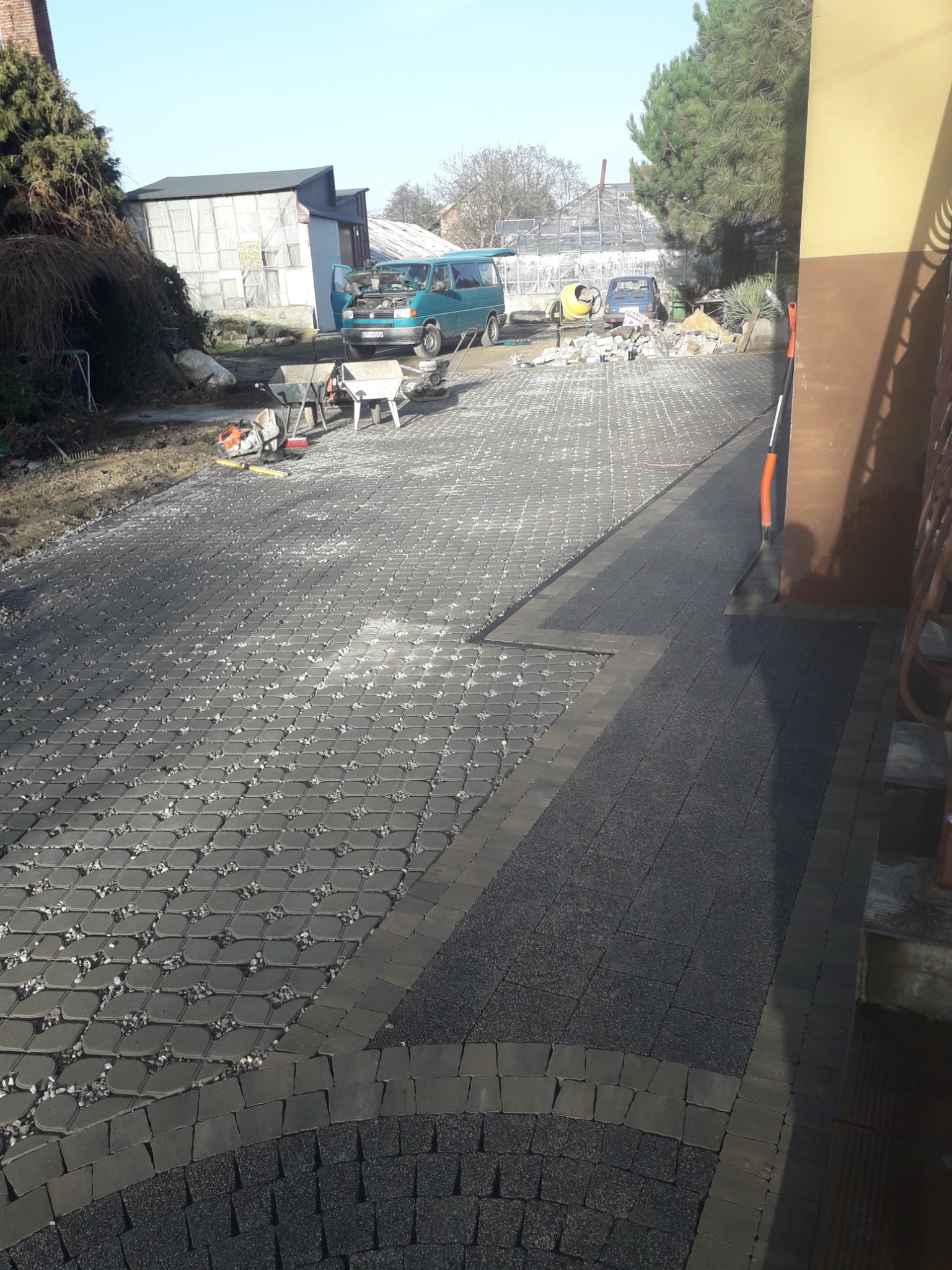 Ułożona nawierzchnia z kostki brukowej w dwóch odcieniach szarości, z widocznymi narzędziami budowlanymi, betoniarką i samochodem dostawczym w tle, sugerująca trwające prace.