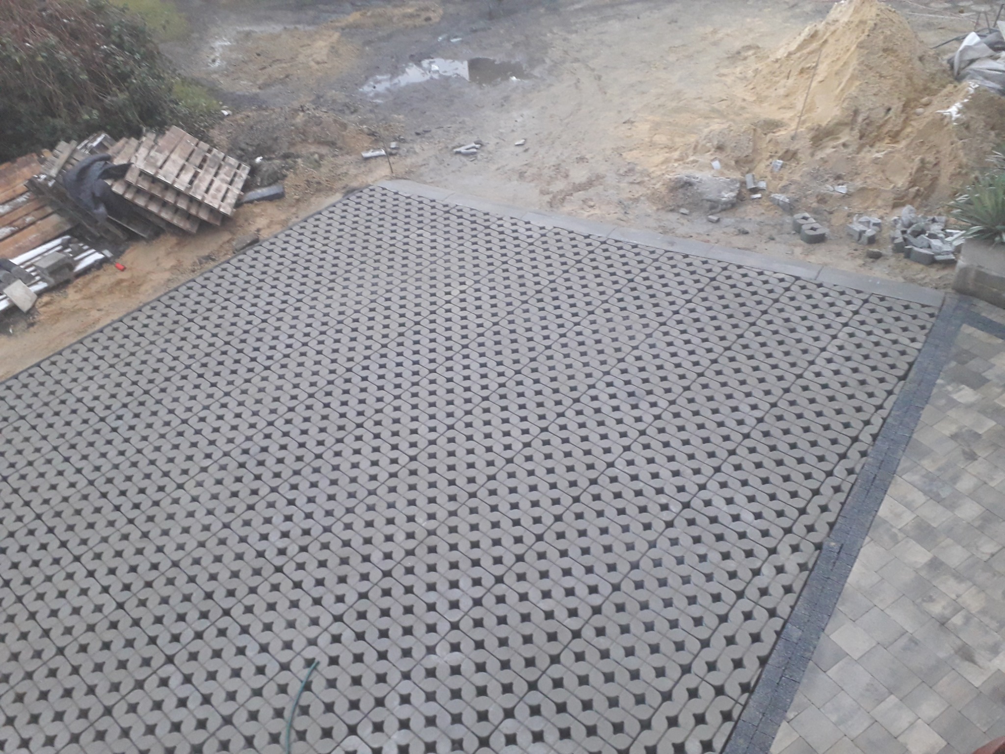 Ułożona nawierzchnia z ażurowych, betonowych płyt z charakterystycznym wzorem, widoczne palety z materiałami budowlanymi i pryzma piasku w tle.