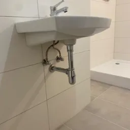 Nowoczesna, biała umywalka ceramiczna zamontowana na ścianie z chromowaną baterią i syfonem, widoczna instalacja wodna i odpływ, w tle kabina prysznicowa.