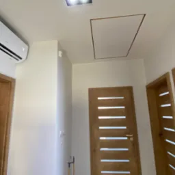Wnętrze korytarza po remoncie z zamontowaną klimatyzacją na ścianie, widoczne trzy drewniane drzwi z przeszkleniami i klapa rewizyjna w suficie.