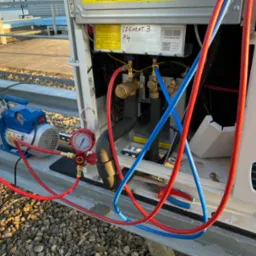 Podłączenie pompy próżniowej do instalacji klimatyzacyjnej na dachu budynku, widoczne manometry, węże w kolorach czerwonym i niebieskim oraz narzędzia.