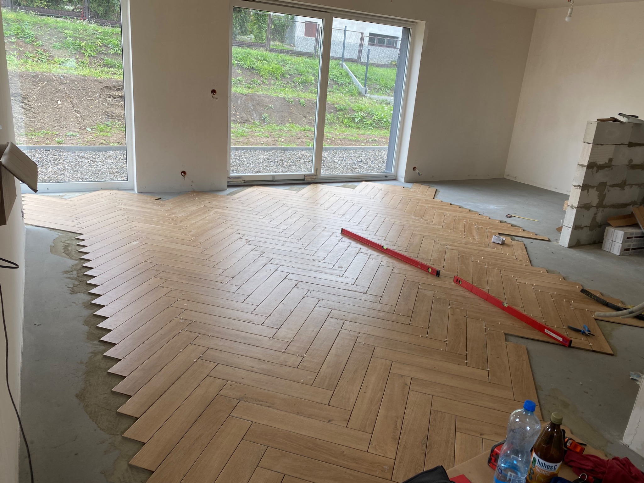 Układanie drewnopodobnej podłogi w jodełkę w jasnym, przestronnym pokoju z dużym oknem przesuwnym; widoczny poziomica, płytki i butelki z napojami.