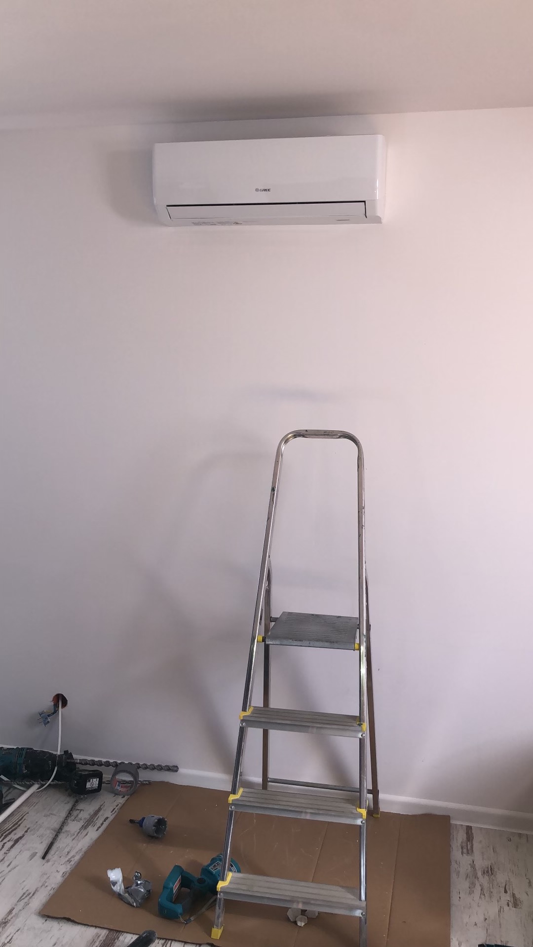 Biały klimatyzator marki Gree zamontowany na ścianie, poniżej rozstawiona aluminiowa drabina, na podłodze karton z narzędziami do montażu.
