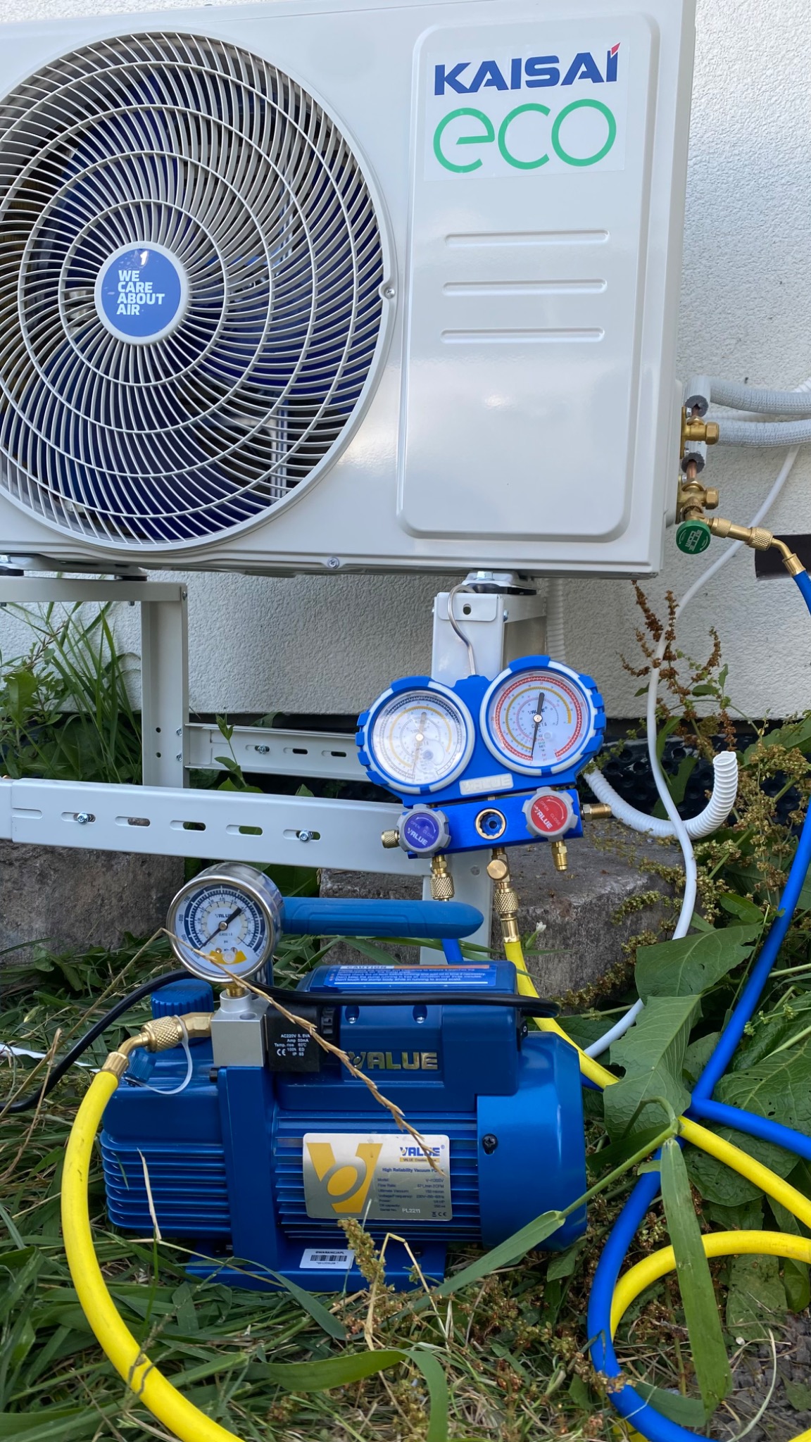 Zewnętrzna jednostka klimatyzacji Kaisai Eco zamontowana na wsporniku, podłączona do manometrów i pompy próżniowej marki Value podczas serwisu na tle zieleni.