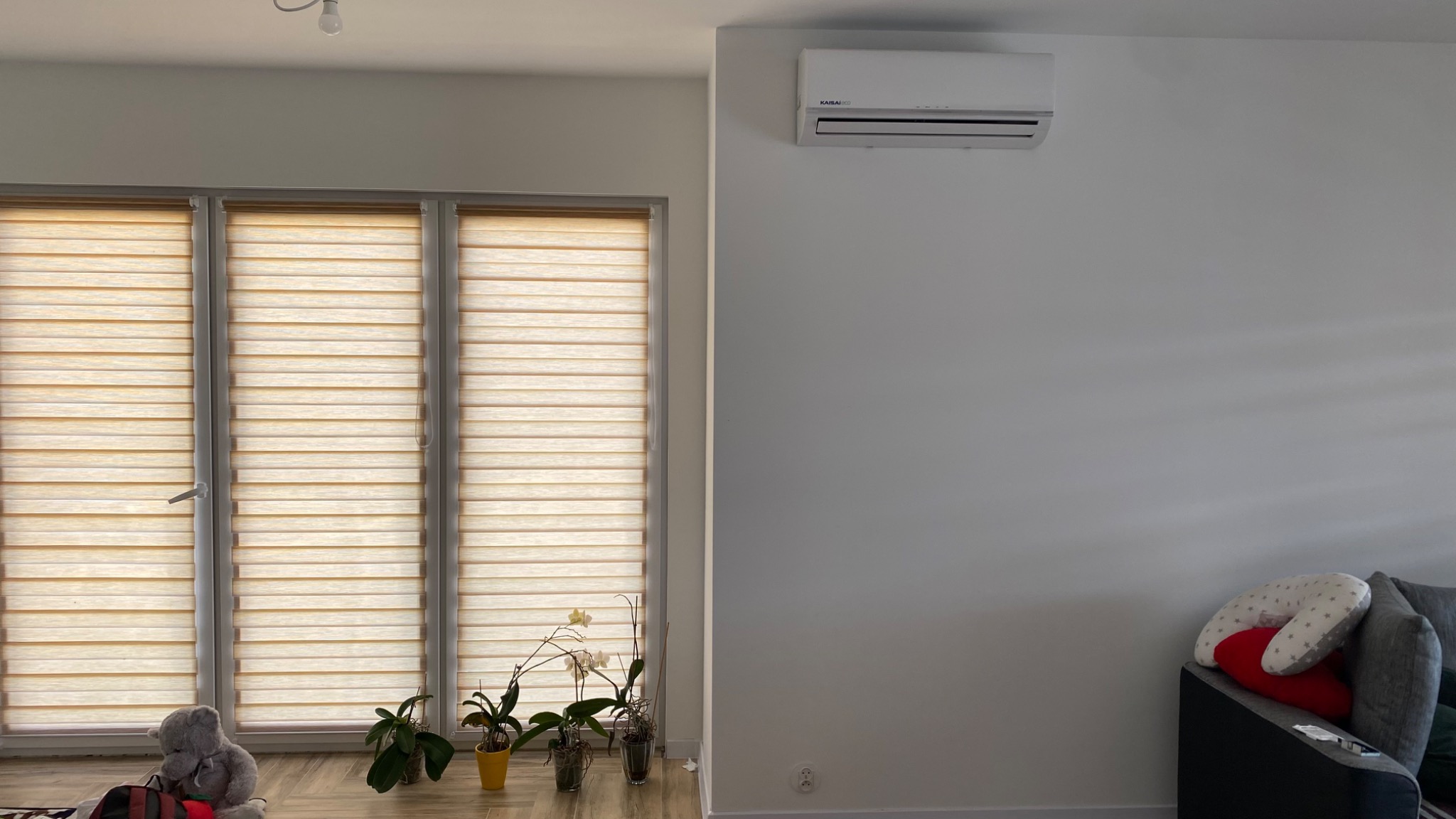 Biały klimatyzator Kaisaico zamontowany na ścianie w jasnym pokoju z trzema oknami z żaluzjami typu dzień i noc, trzema orchideami w doniczkach oraz szarą kanapą z poduszkami.