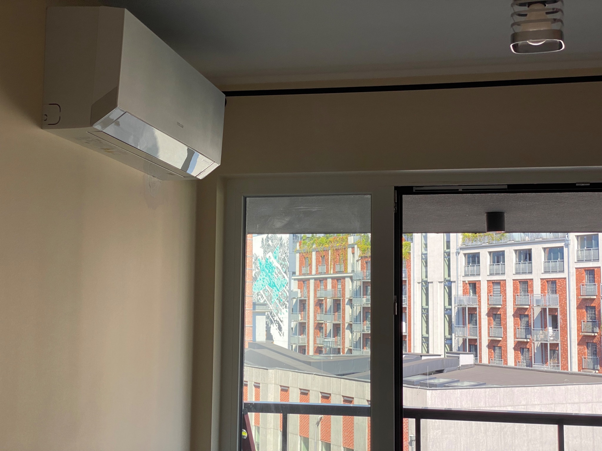 Biała klimatyzacja zamontowana na ścianie w mieszkaniu z widokiem na nowoczesne budynki za oknem.