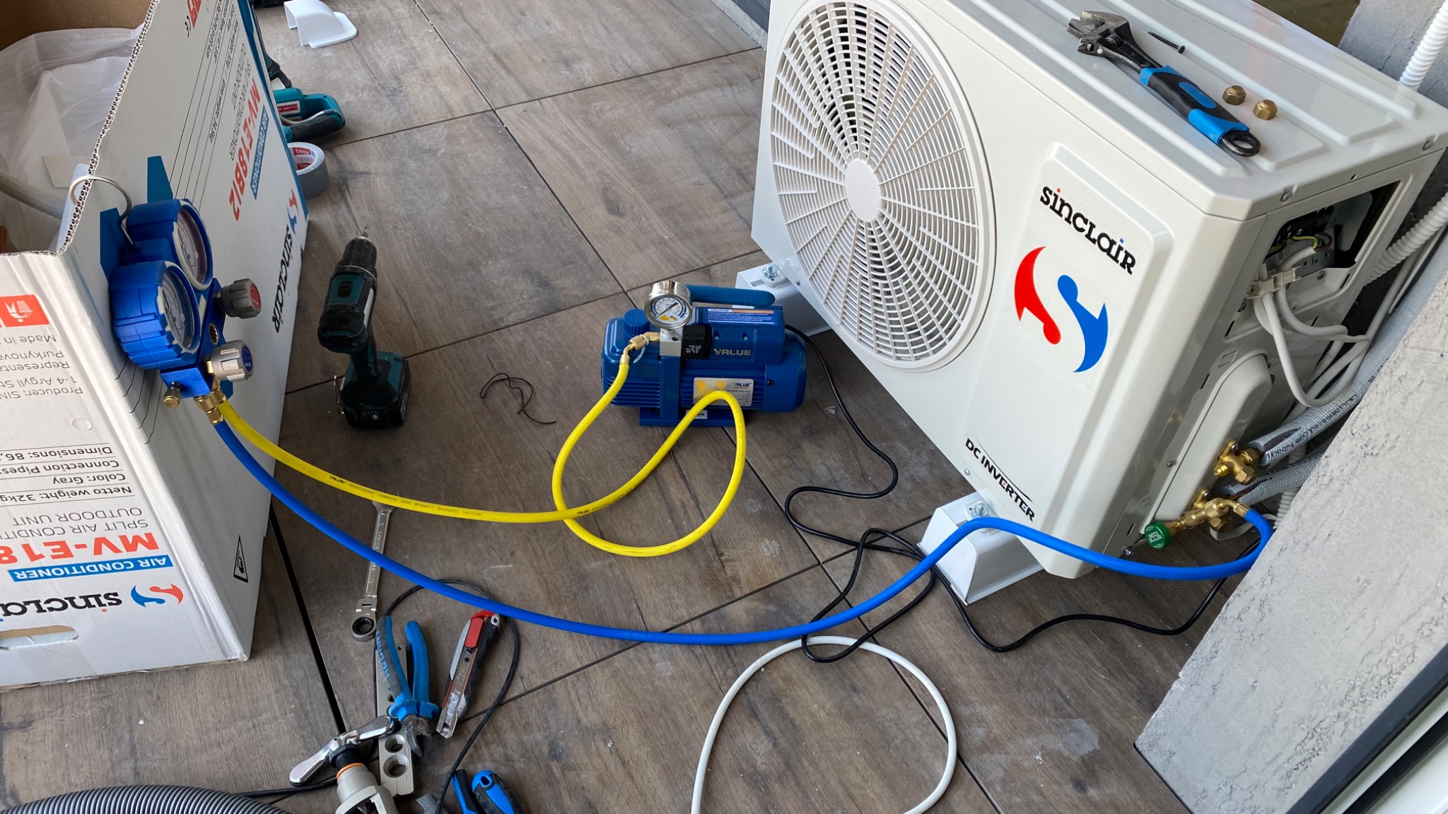 Instalacja zewnętrznej jednostki klimatyzacji Sinclair z podłączoną pompą próżniową Value i zestawem manometrów, widoczne narzędzia: wkrętarka, klucze, kombinerki, przewody chłodnicze.