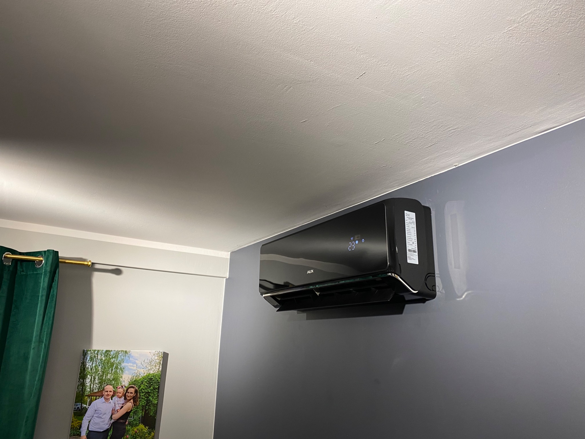 Czarny klimatyzator AUX zamontowany na szarej ścianie w pokoju. Na ścianie wisi również obraz z rodziną, a w rogu widać zieloną zasłonę.