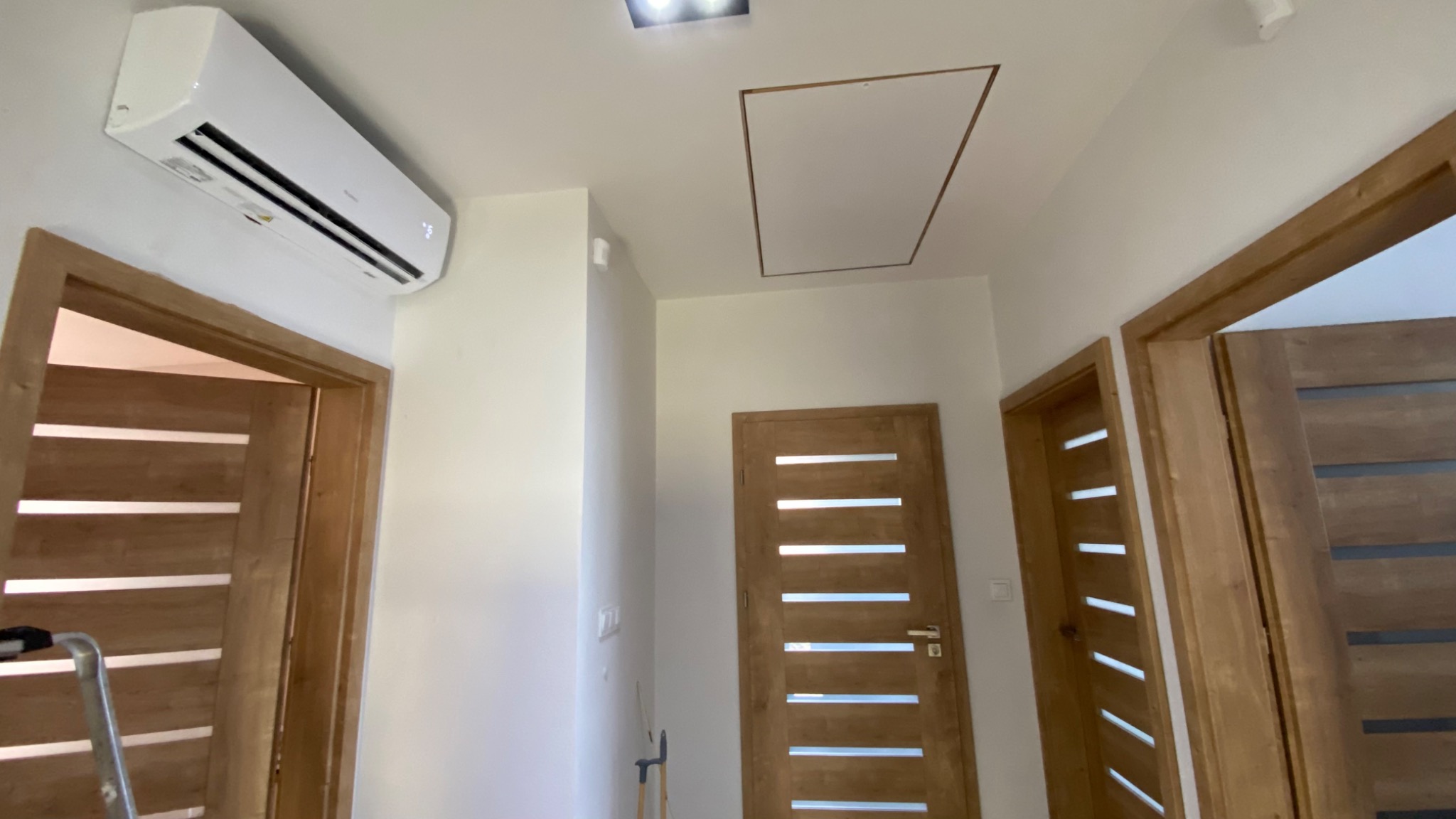 Wnętrze korytarza po remoncie z zamontowaną klimatyzacją na ścianie, widoczne trzy drewniane drzwi z przeszkleniami i klapa rewizyjna w suficie.