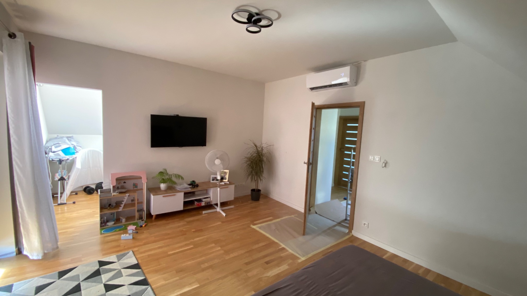Jasne wnętrze pokoju na poddaszu z klimatyzatorem zamontowanym na ścianie nad drzwiami, widoczny telewizor, zabawki i wentylator stojący.