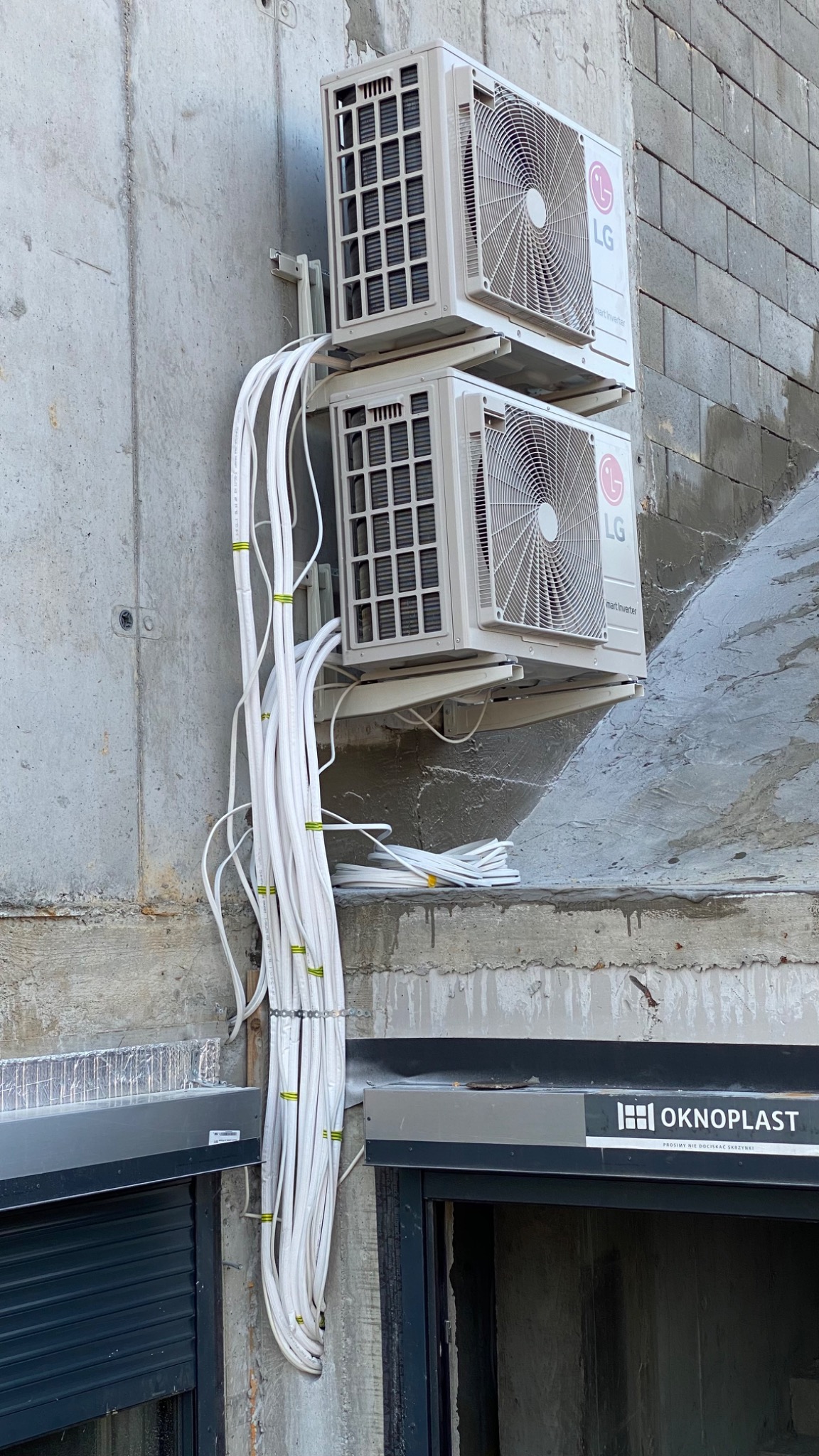 Dwa agregaty klimatyzacyjne LG zamontowane na szarej ścianie budynku, widoczne wiązki białych kabli z żółtymi opaskami. Pod spodem okno z roletami i logo Okoplast.