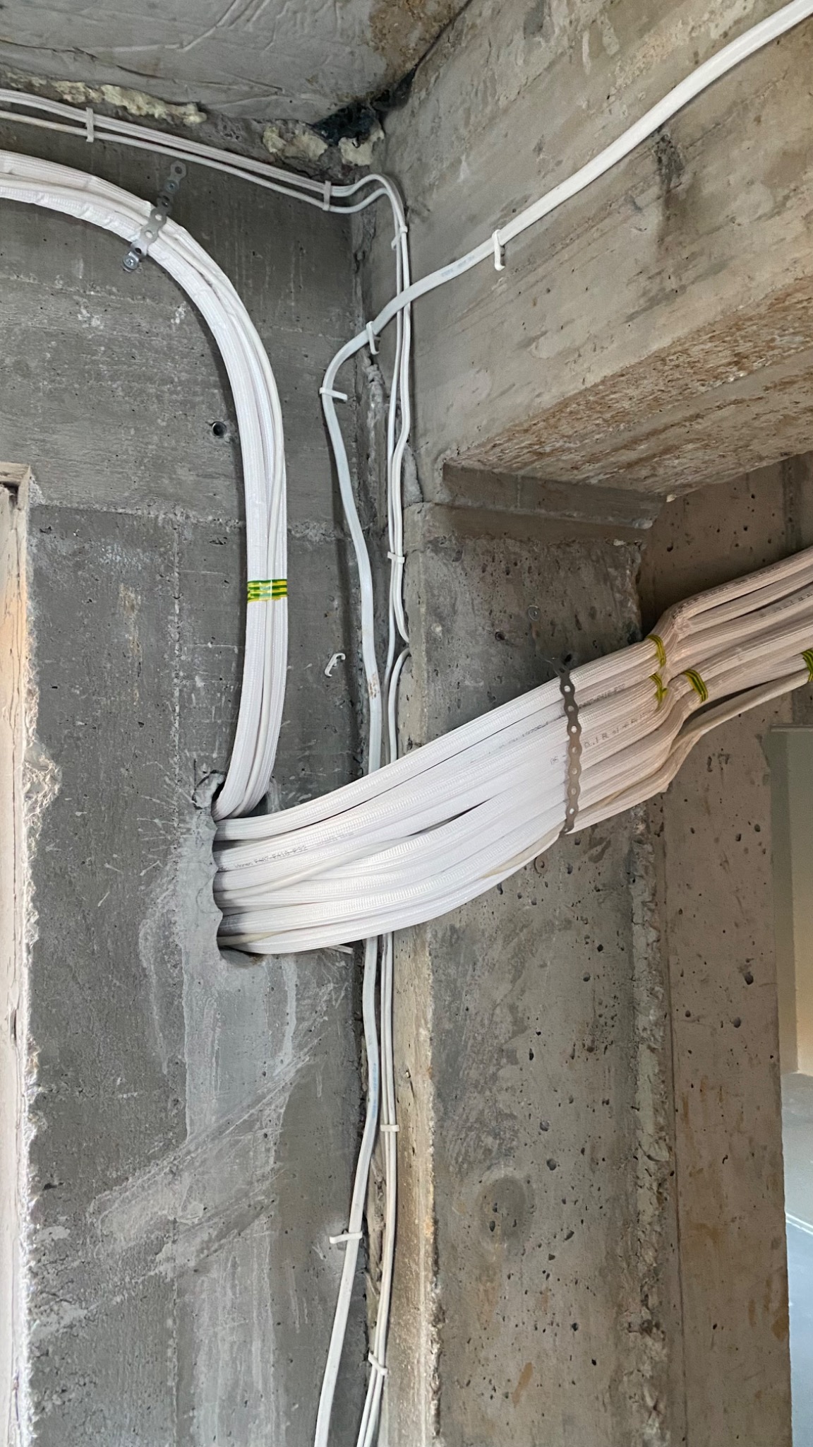 Białe rury instalacyjne, zabezpieczone zielonymi taśmami i metalowymi uchwytami, prowadzone wzdłuż surowej, betonowej ściany w nowym budynku.