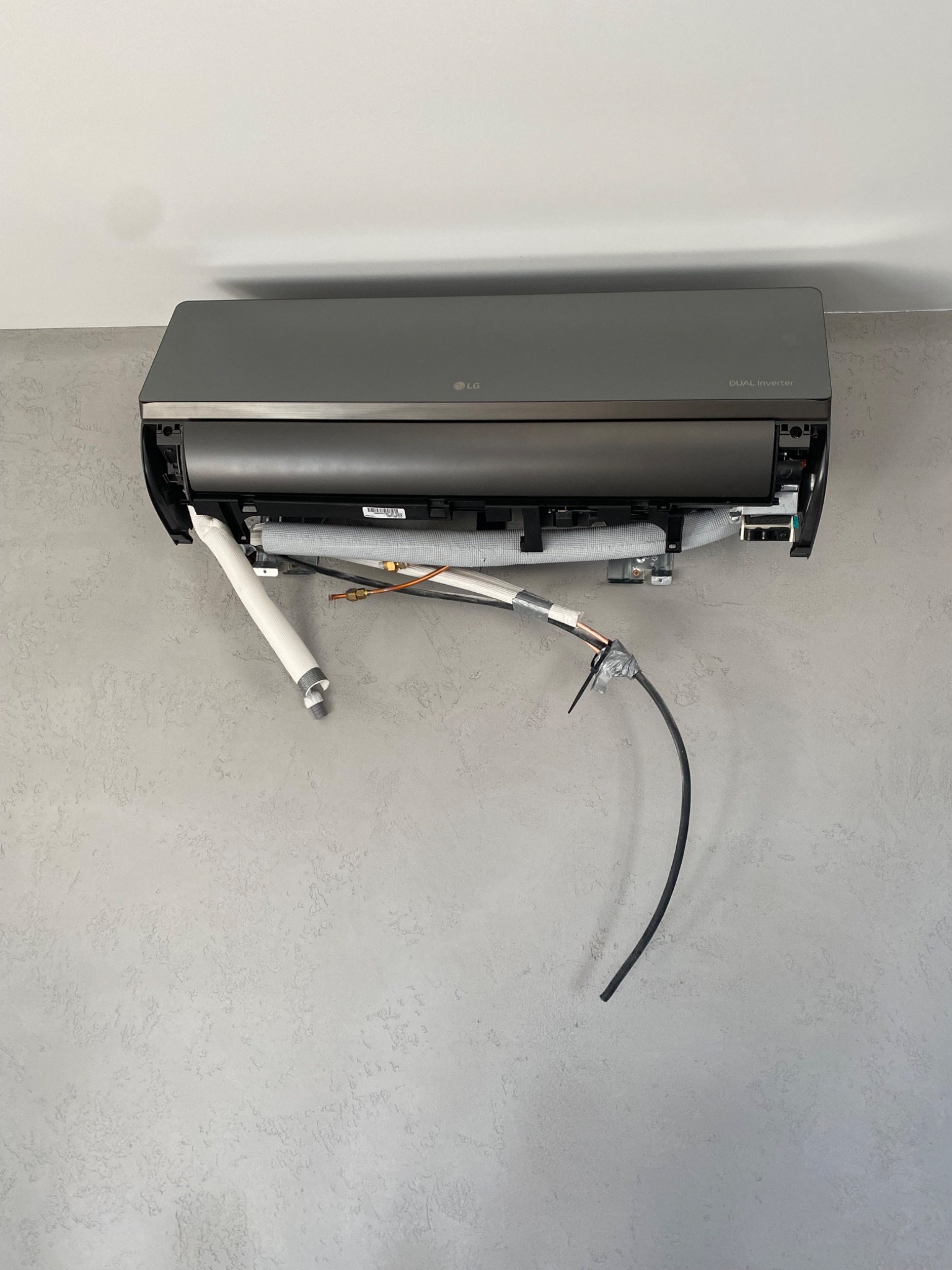 Szary klimatyzator LG Dual Inverter zamontowany na ścianie z widocznymi przewodami i rurami instalacyjnymi w trakcie montażu.