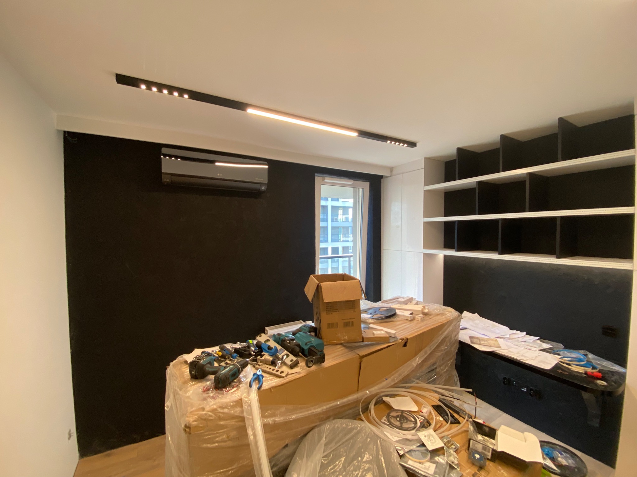 Wnętrze w trakcie wykańczania: zamontowana klimatyzacja, oświetlenie liniowe LED, regał z czarnymi półkami, narzędzia i materiały budowlane na zabezpieczonym folią blacie.
