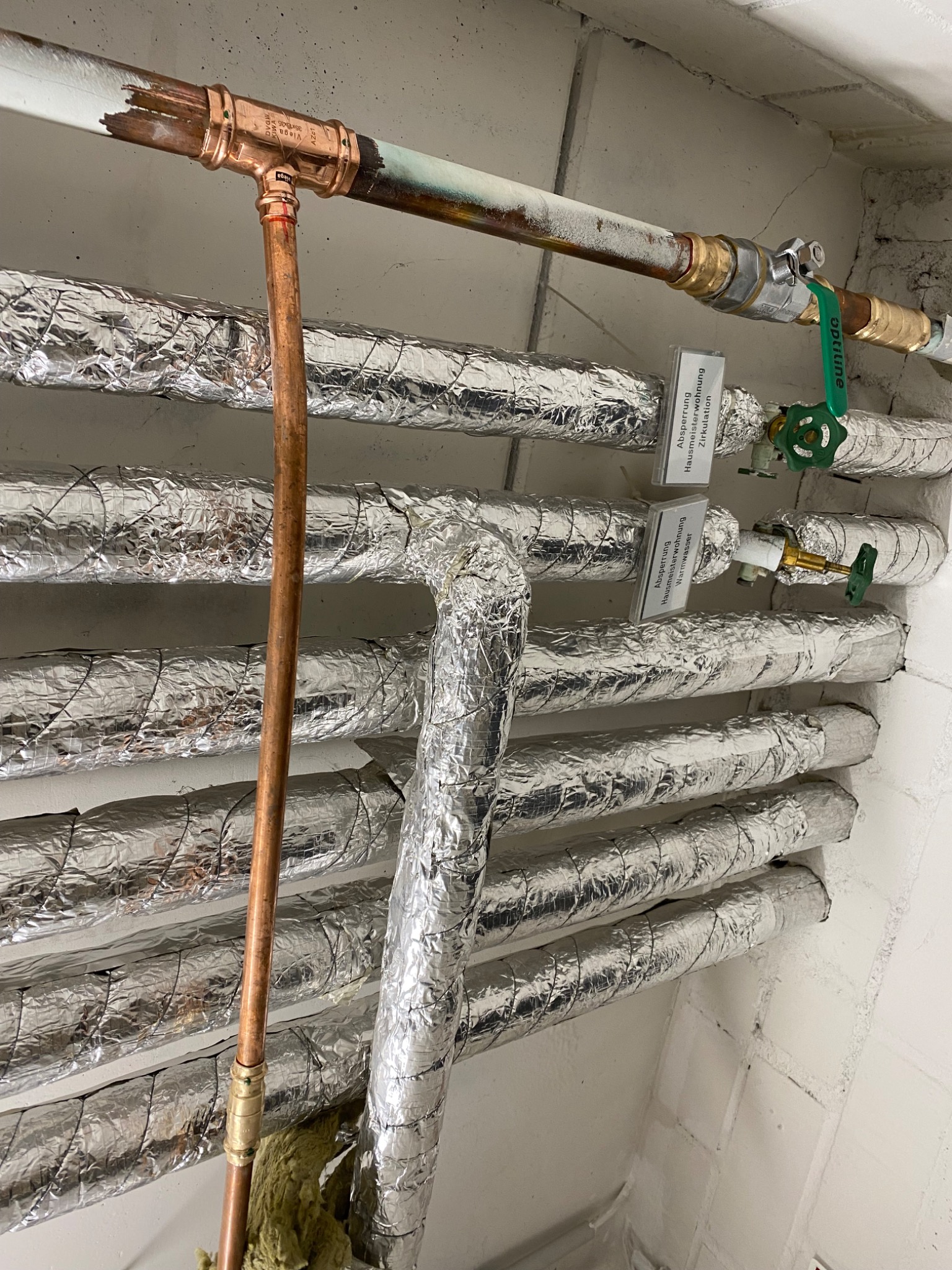 Rzędy rur centralnego ogrzewania owiniętych srebrną folią izolacyjną, z zaworami odcinającymi i etykietami w języku niemieckim, oraz pionowa rura miedziana z nowym złączem.