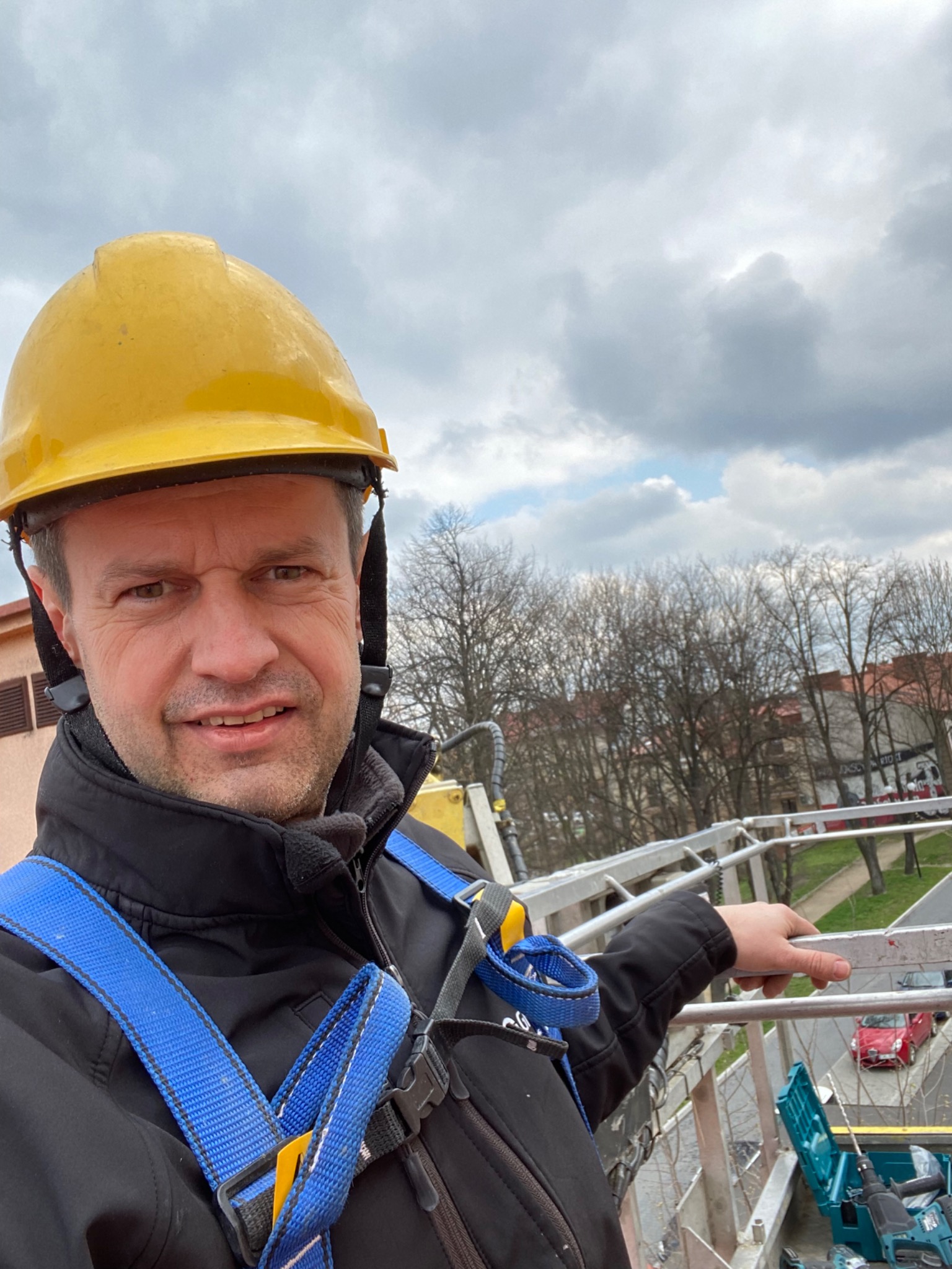 Pracownik w żółtym kasku i uprzęży zabezpieczającej na wysokości, wskazujący na zewnątrz, z widocznym sprzętem i narzędziami w tle.