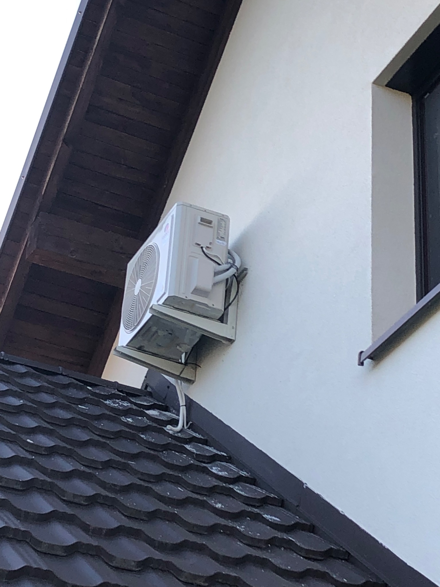Biała jednostka zewnętrzna klimatyzatora zamontowana na elewacji domu, widoczna z perspektywy dachu pokrytego ciemną dachówką. Zewnętrzne kable i rury prowadzone wzdłuż ściany.