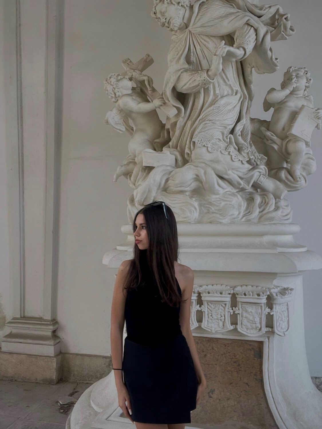 Młoda kobieta w czarnej sukience pozuje na tle białej, rzeźbionej kolumny z figurami aniołów i świętego, spoglądając w lewo.