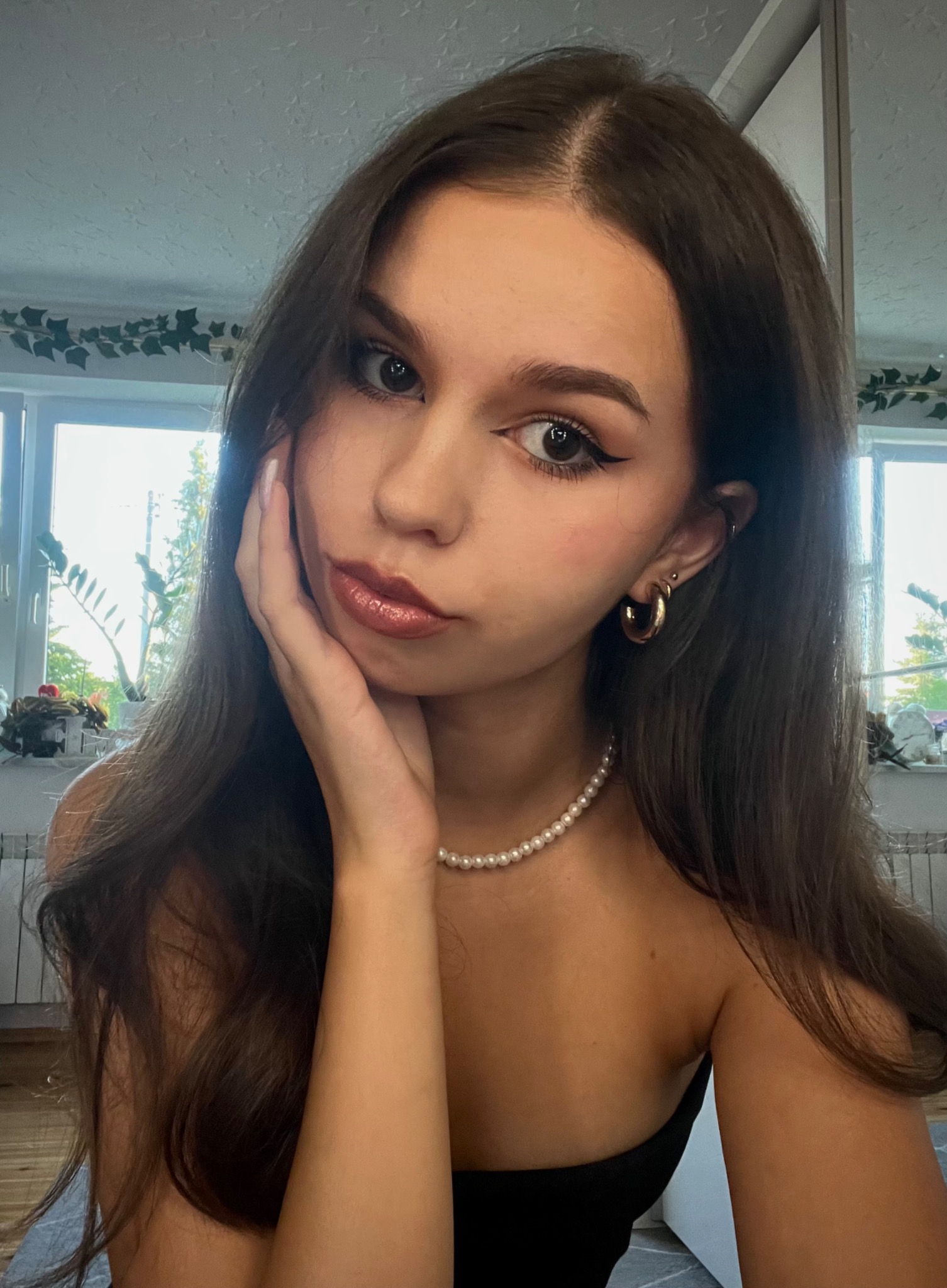 Portret młodej kobiety z długimi brązowymi włosami, delikatnym makijażem, perłowym naszyjnikiem i złotymi kolczykami na tle okna z roślinami.