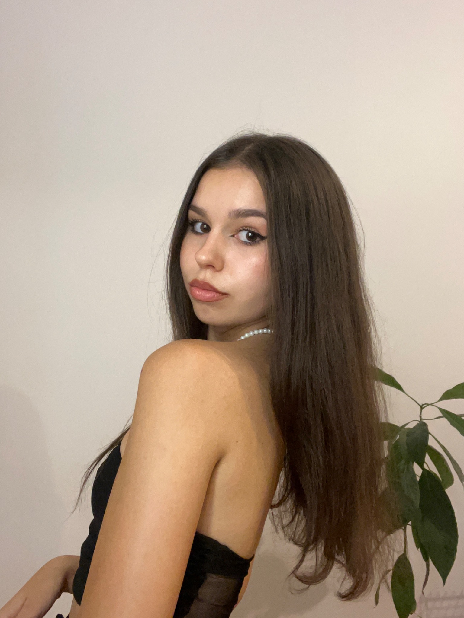 Portret młodej kobiety z długimi, brązowymi włosami, spoglądającej przez ramię, z delikatnym makijażem i perłowym naszyjnikiem, na neutralnym tle z rośliną.