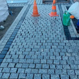 Świeżo ułożona nawierzchnia z kostki granitowej, obramowana ciemniejszymi kostkami, z pomarańczowymi pachołkami i pracownikiem polewającym wodą, w tle palety z materiałami budowlanymi.