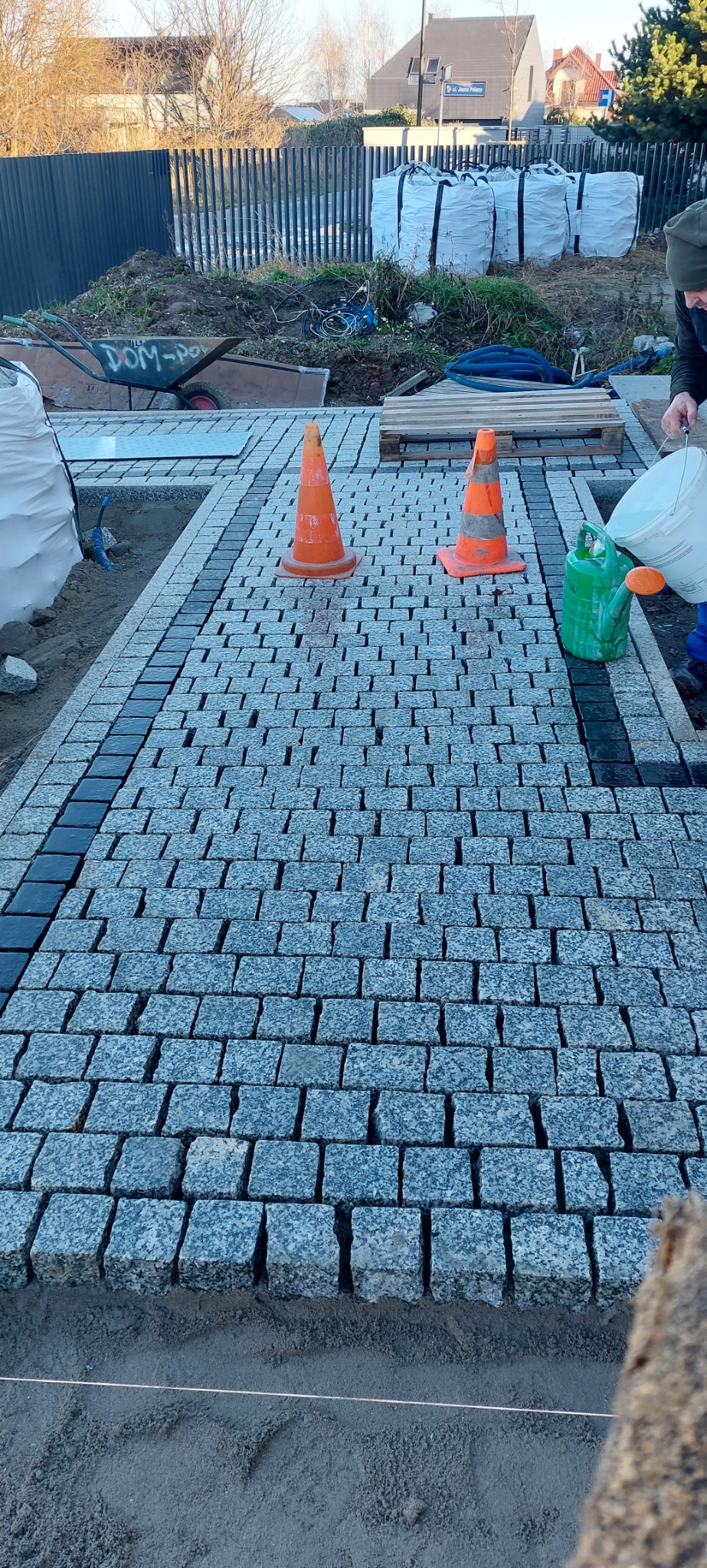 Świeżo ułożona nawierzchnia z kostki granitowej, obramowana ciemniejszymi kostkami, z pomarańczowymi pachołkami i pracownikiem polewającym wodą, w tle palety z materiałami budowlanymi.