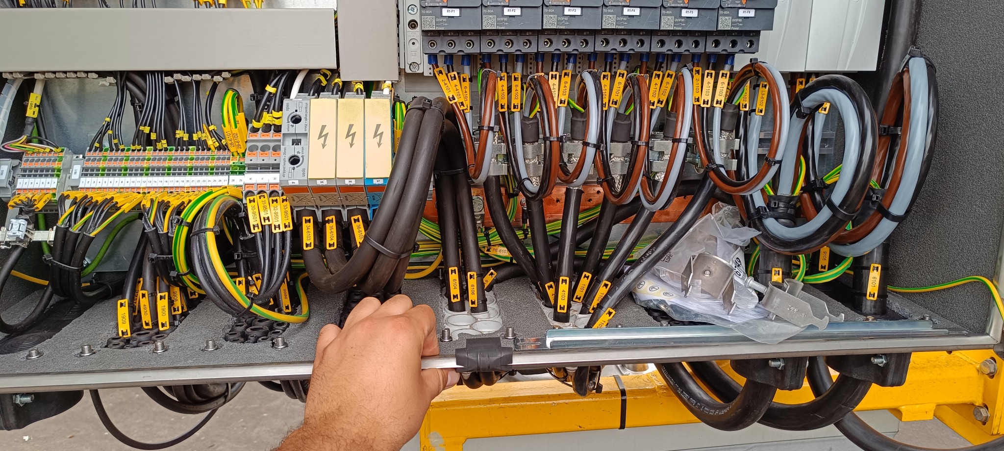 Otwarta szafa elektryczna w trakcie instalacji, widoczne liczne przewody oznaczone żółtymi etykietami, listwy zaciskowe, moduły zabezpieczeń i dłoń elektryka przytrzymująca element konstrukcyjny.