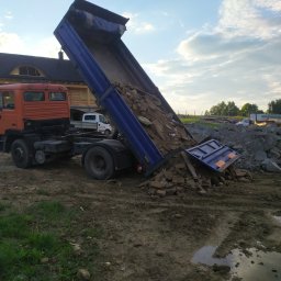 STOBart - Pomarańczowa wywrotka z podniesioną skrzynią, wysypująca gruz na błotniste podłoże. W tle drewniany dom i zachmurzone niebo.