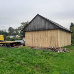 Nowo wybudowany drewniany budynek gospodarczy z żółtą lawetą transportową w tle, na trawniku leżą resztki cegieł i desek.