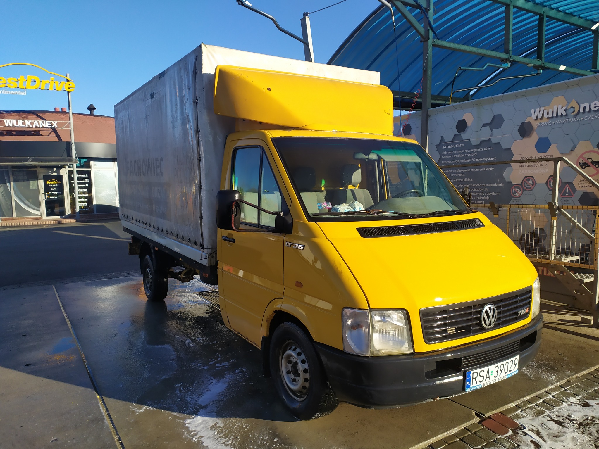 Żółty samochód dostawczy LT35 z plandeką, zaparkowany na mokrym placu przed budynkiem Wulkanex, w słoneczny dzień. Widoczna tablica rejestracyjna.