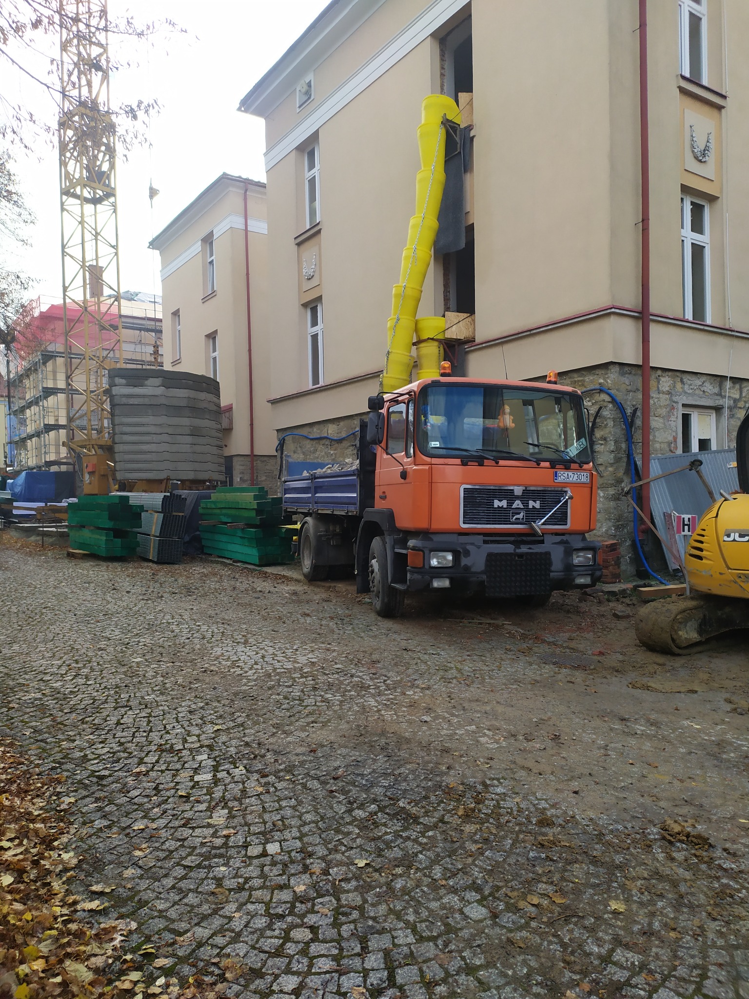Pomarańczowa ciężarówka MAN z żółtym rękawem zsypowym przy budynku w trakcie rozbiórki. W tle widoczny dźwig i materiały budowlane.