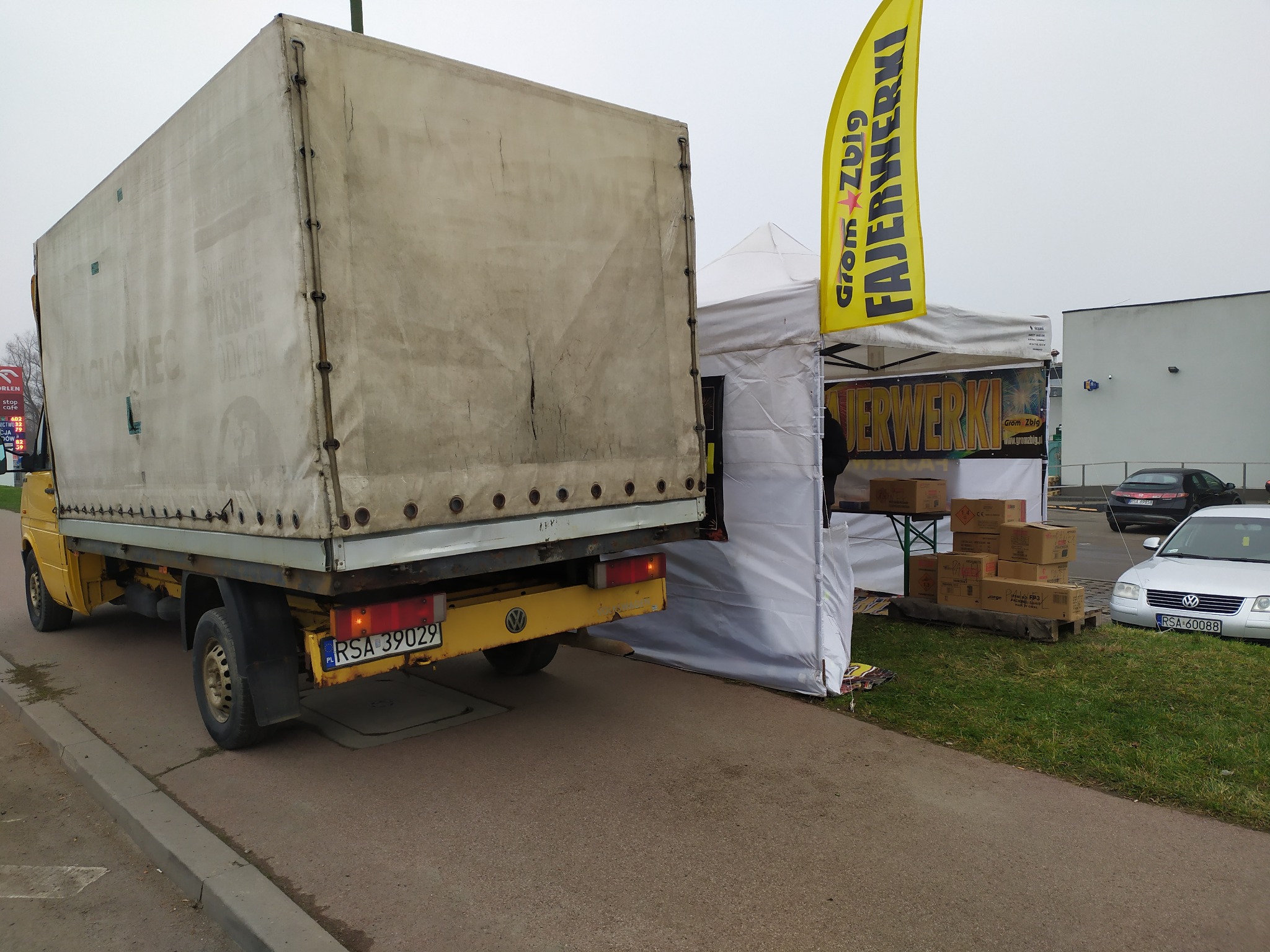 Żółty samochód dostawczy z plandeką zaparkowany obok stoiska z fajerwerkami, widoczny fragment żółtej flagi reklamowej i kartony z produktami.