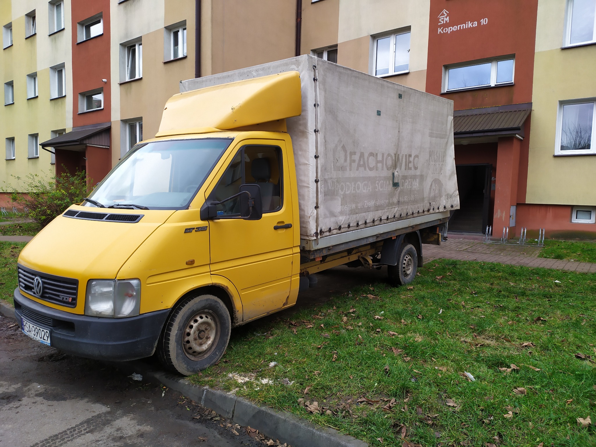 Żółty samochód dostawczy z plandeką firmy transportowej 'Fachowiec' zaparkowany przed budynkiem mieszkalnym przy ulicy Kopernika 10. Widoczny numer rejestracyjny.