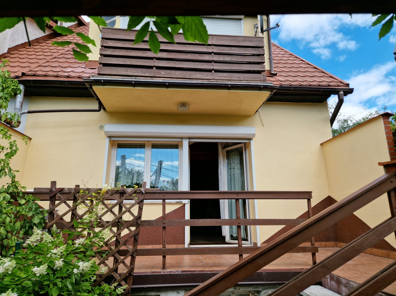 Żółta elewacja domu jednorodzinnego z drewnianym balkonem i schodami, widoczne okno z roletą i otwarte drzwi balkonowe, częściowo przysłonięte przez zieloną roślinność.