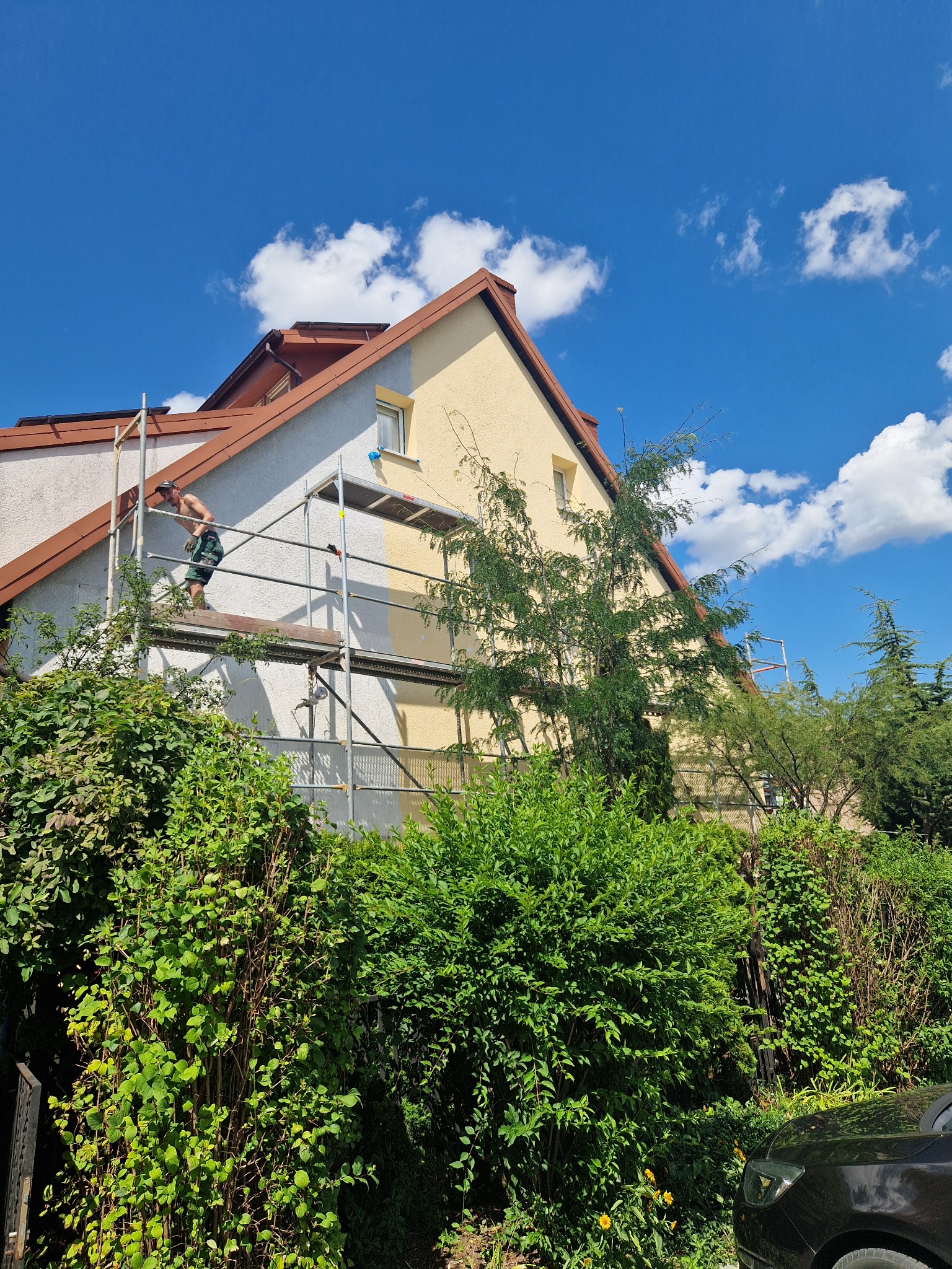 Pracownik na rusztowaniu maluje elewację domu na tle błękitnego nieba z białymi chmurami; fragment ściany w kolorze żółtym, a fragment w kolorze szarym.