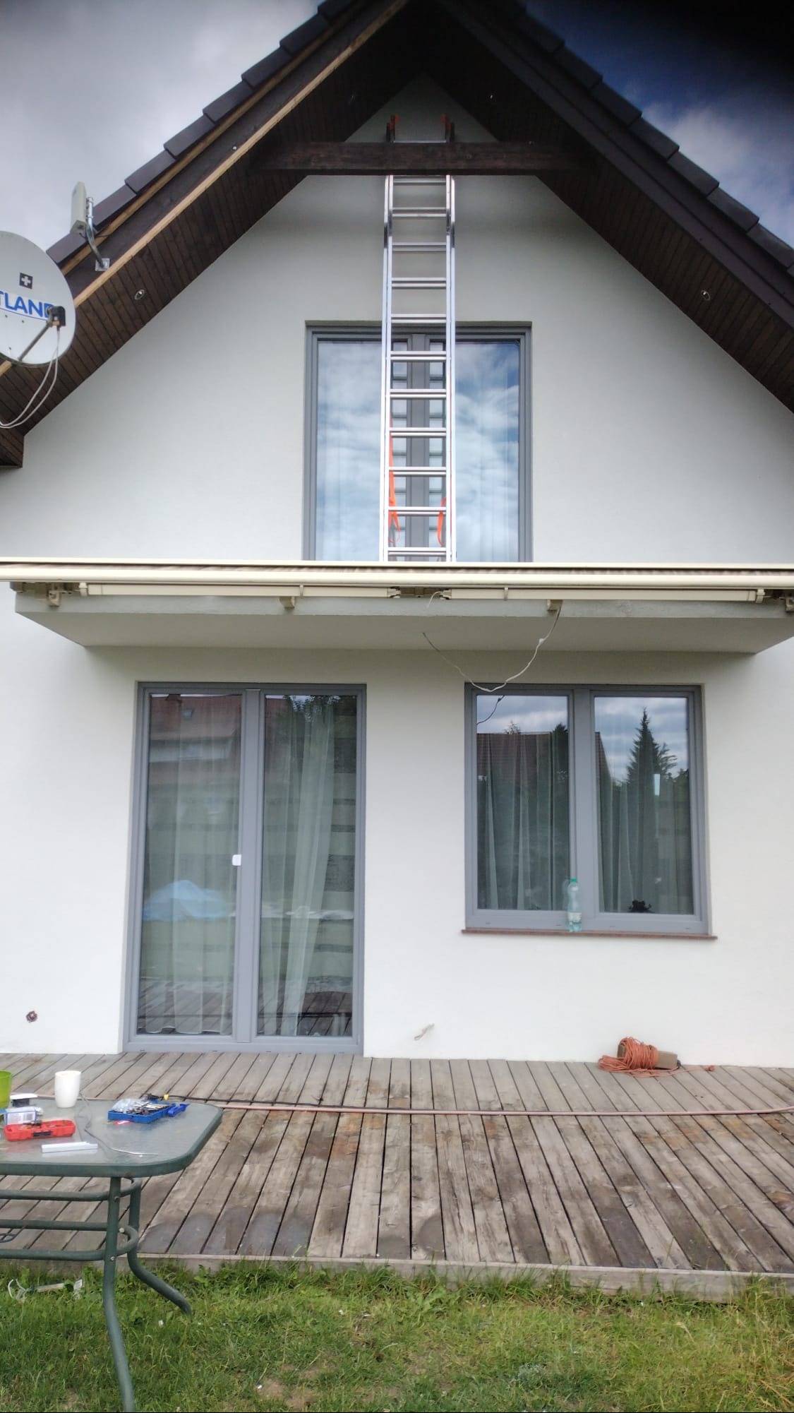 Dwukondygnacyjny dom z jasną elewacją, drewnianym dachem i aluminiową drabiną opartą o okno na poddaszu, widok z zewnątrz na drewniany taras z narzędziami i butelką wody.