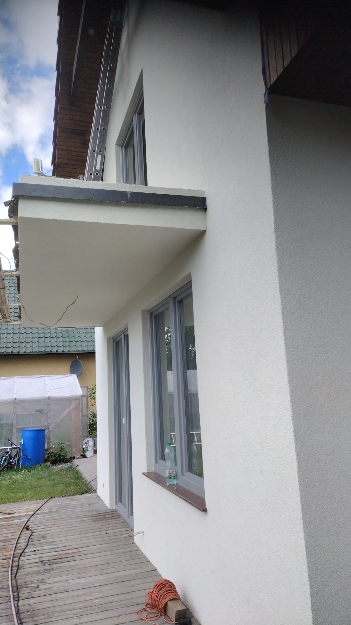 Świeżo otynkowana, biała elewacja domu jednorodzinnego z szarymi ramami okiennymi i balkonem, widoczna drabina oparta o dach, butelka wody na parapecie, drewniany taras i zwinięty kabel.