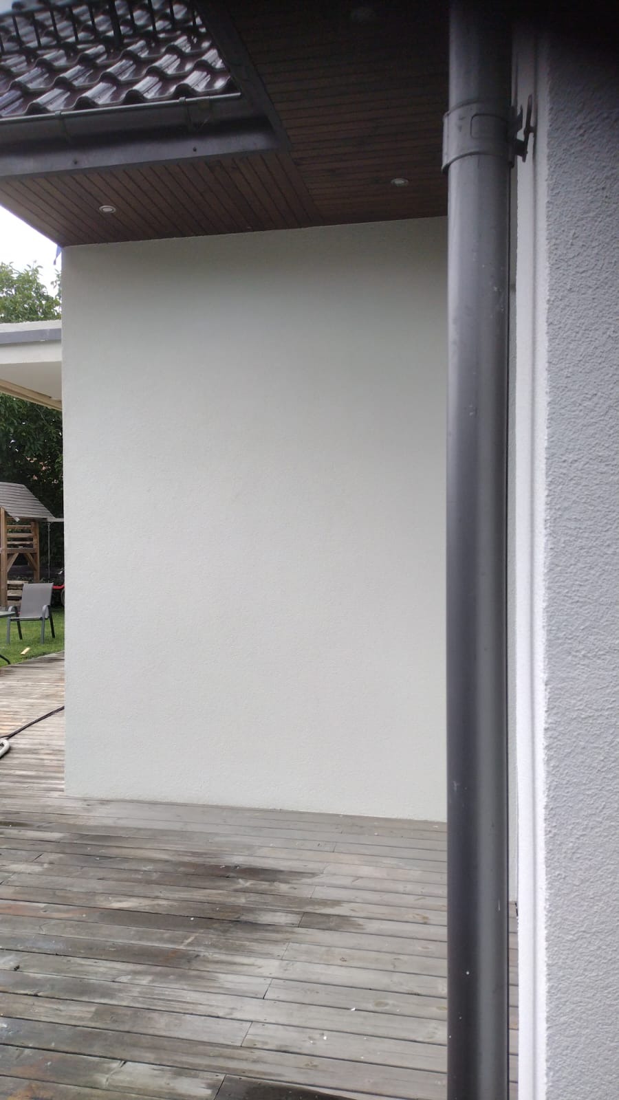 Fragment elewacji domu z widoczną rynną spustową, świeżo otynkowaną ścianą i drewnianym tarasem. W tle widoczny plac zabaw.