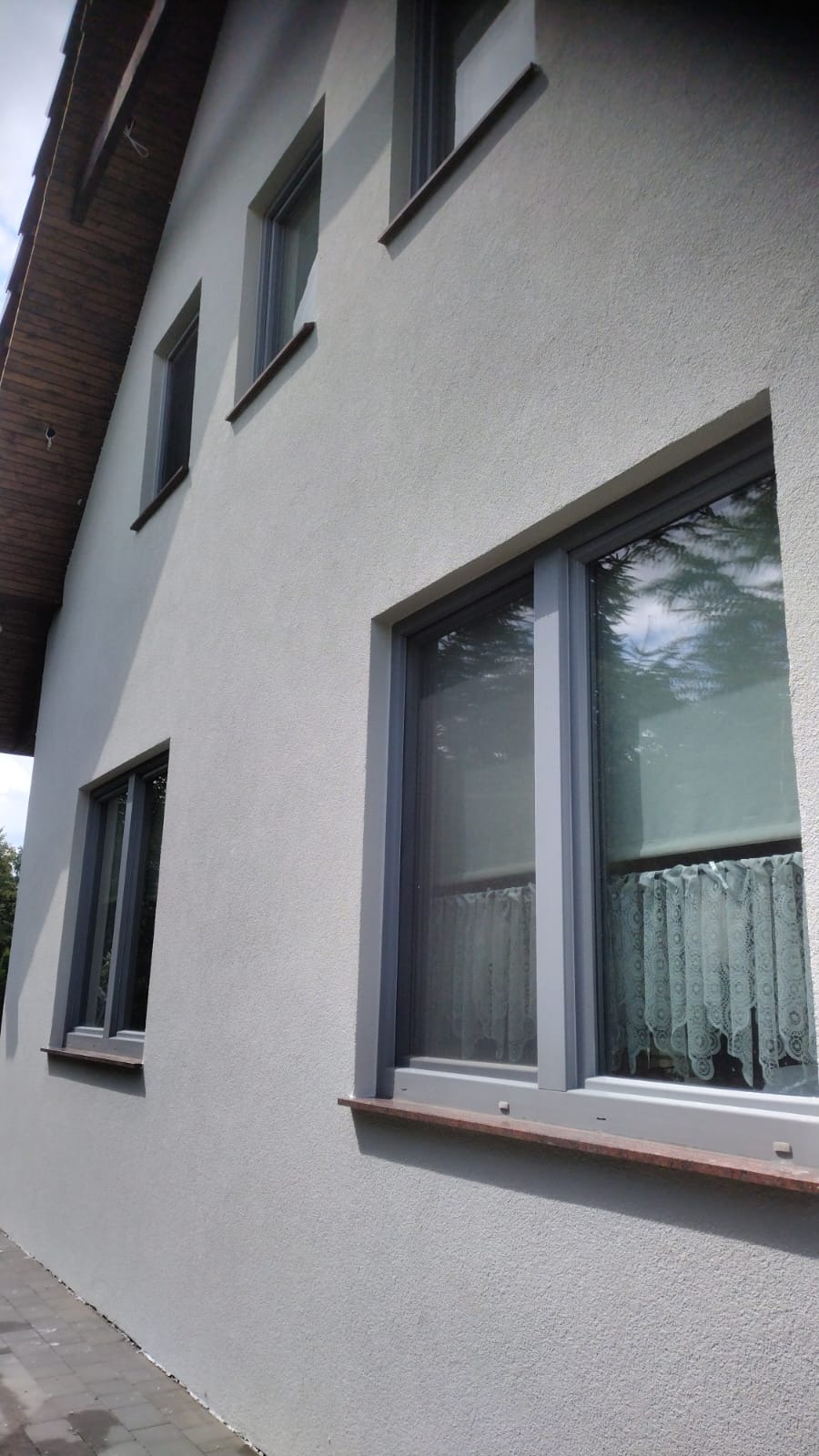 Ściana domu po ociepleniu i nałożeniu jasnoszarego tynku strukturalnego, okna z szarymi ramami i brązowymi parapetami, widok z dołu.