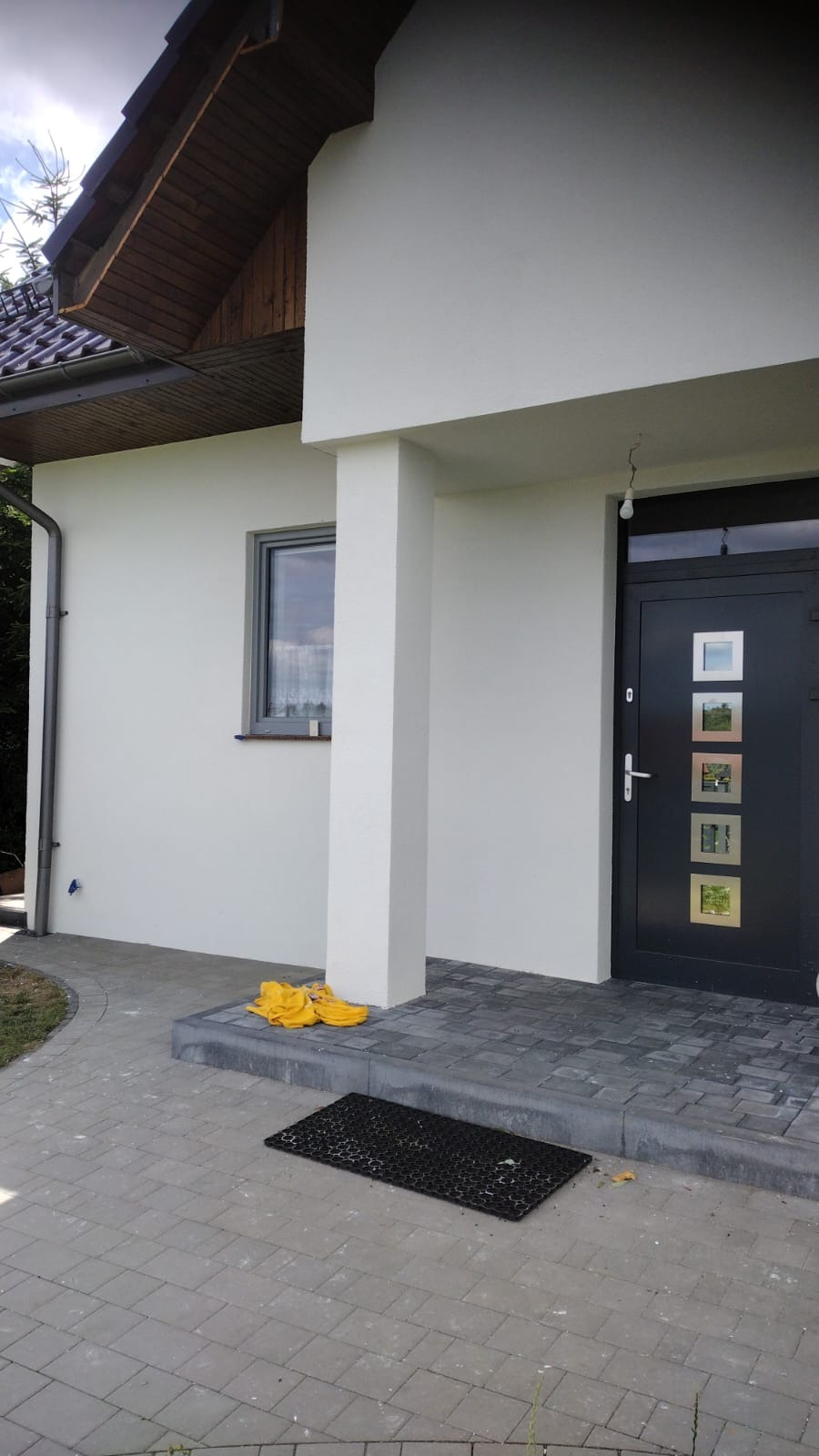 Wykończona elewacja domu jednorodzinnego z czarnymi drzwiami wejściowymi z kwadratowymi przeszkleniami, widoczny fragment chodnika z kostki brukowej i żółte rękawice robocze leżące obok wejścia.